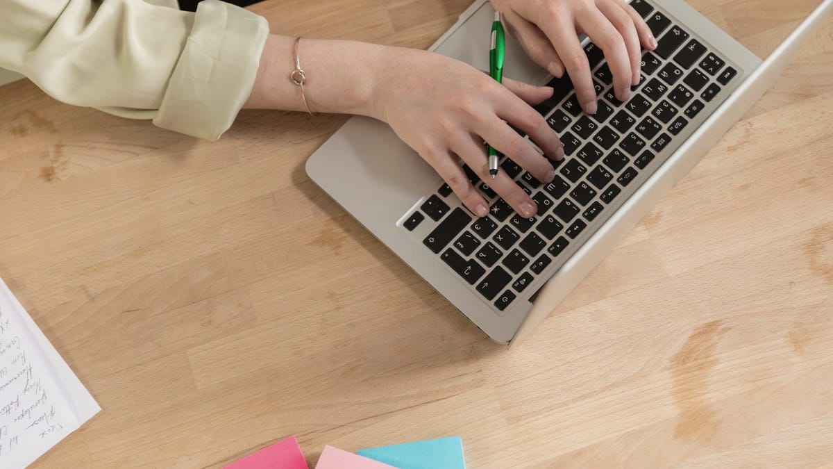 Hands type on a laptop on a wooden desk with a green pen and sticky notes nearby.