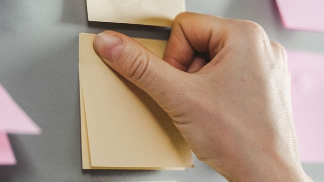 A hand places a blank sticky note on a gray wall with other notes nearby.