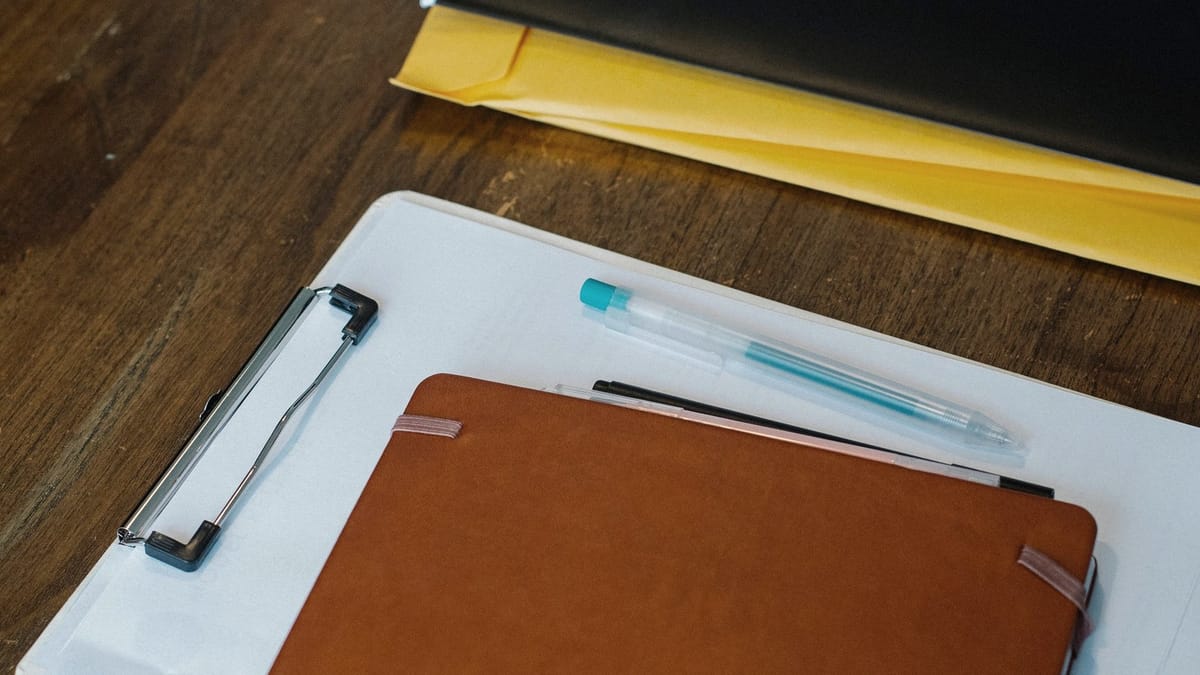 A clipboard, notebook, folder, and pen arranged on a wooden desk.
