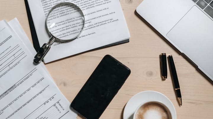 Documents, a magnifying glass, pens, a phone, and a laptop on a desk.